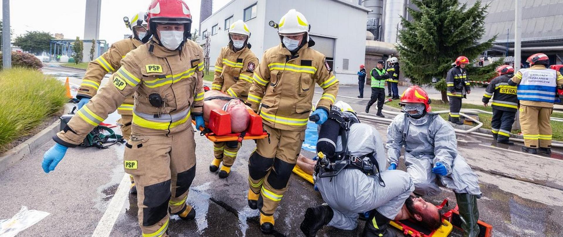 Na zdjęciu widzimy strażaków ubranych w umundurowanie specjalne, którzy niosą na noszach osobę poszkodowaną, a także dwóch strażaków z specjalistycznej grupy ratownictwa chemicznego, którzy udzielają pomocy innej osobie poszkodowanej. W tle widoczni są inni strażacy, a także obiekty huty szkła.