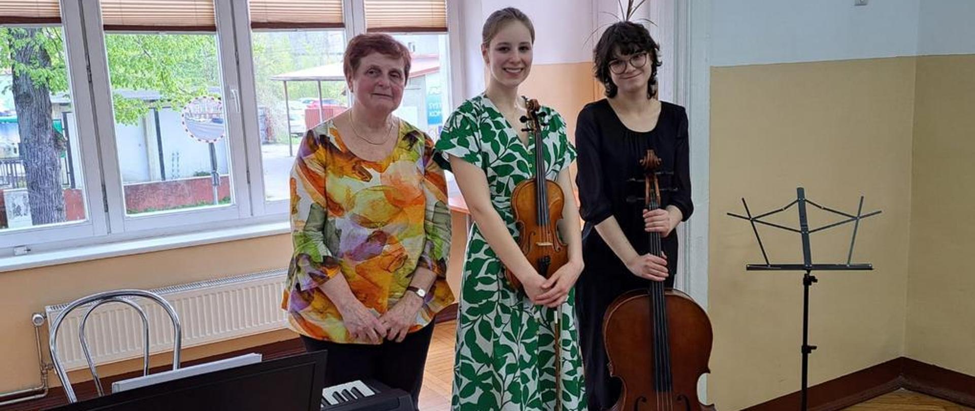 Zdjęcie kolorowe. Uczennice wraz z akompaniatorką kłaniają się po koncercie. Po lewej stronie cyfrowy fortepian. W tle ulica.