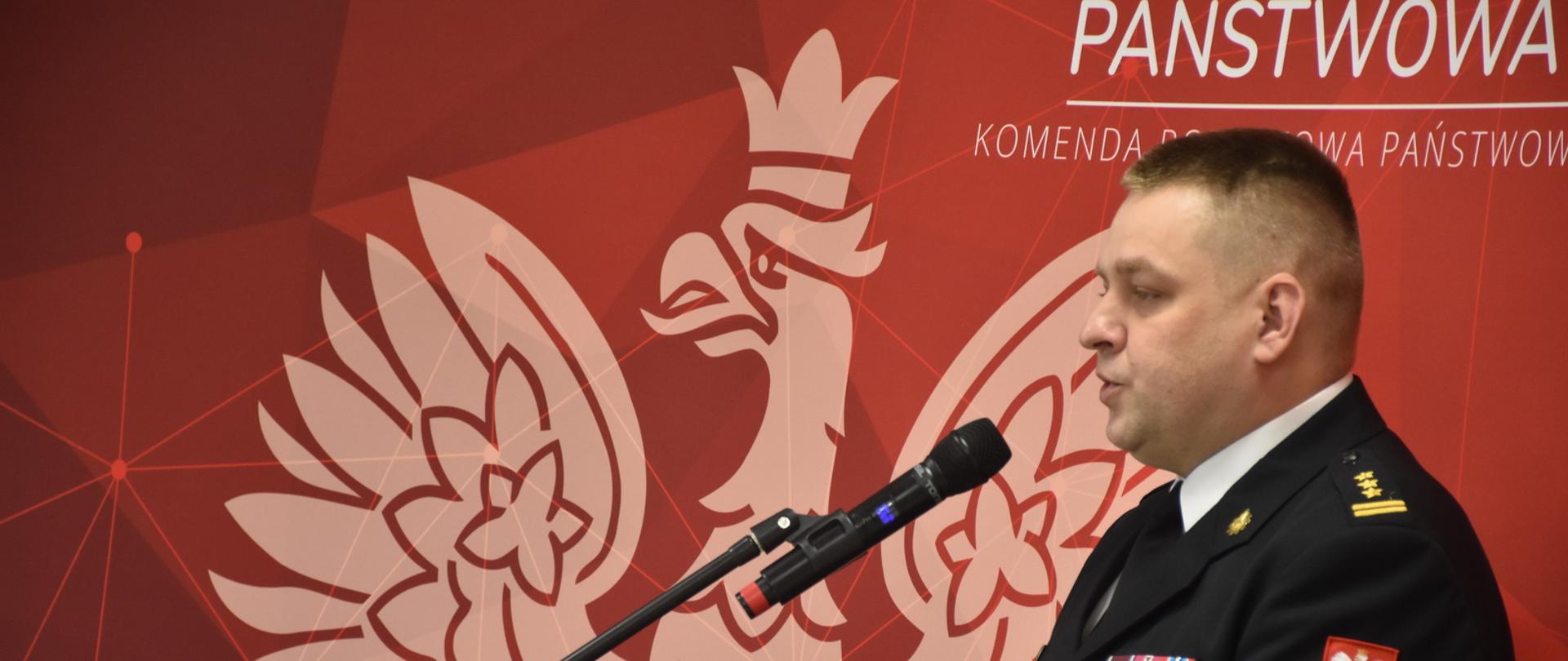 Mężczyzna w mundurze wyjściowym państwowej straży pożarnej w stopniu starszego brygadiera stoi za mównicą i przemawia do mikrofonu, widać jego popiersie, a w tle banner z Godłem Polski