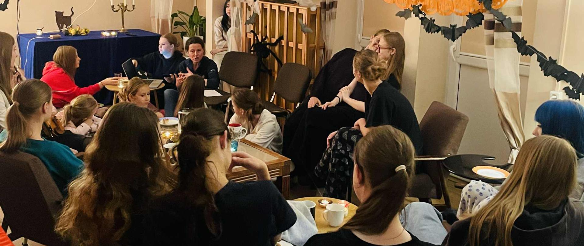 Na zdjęciu grupa dziewcząt i wychowawczyni. Wszyscy siedzą przy stolikach, rozmawiają i uśmiechają się do siebie