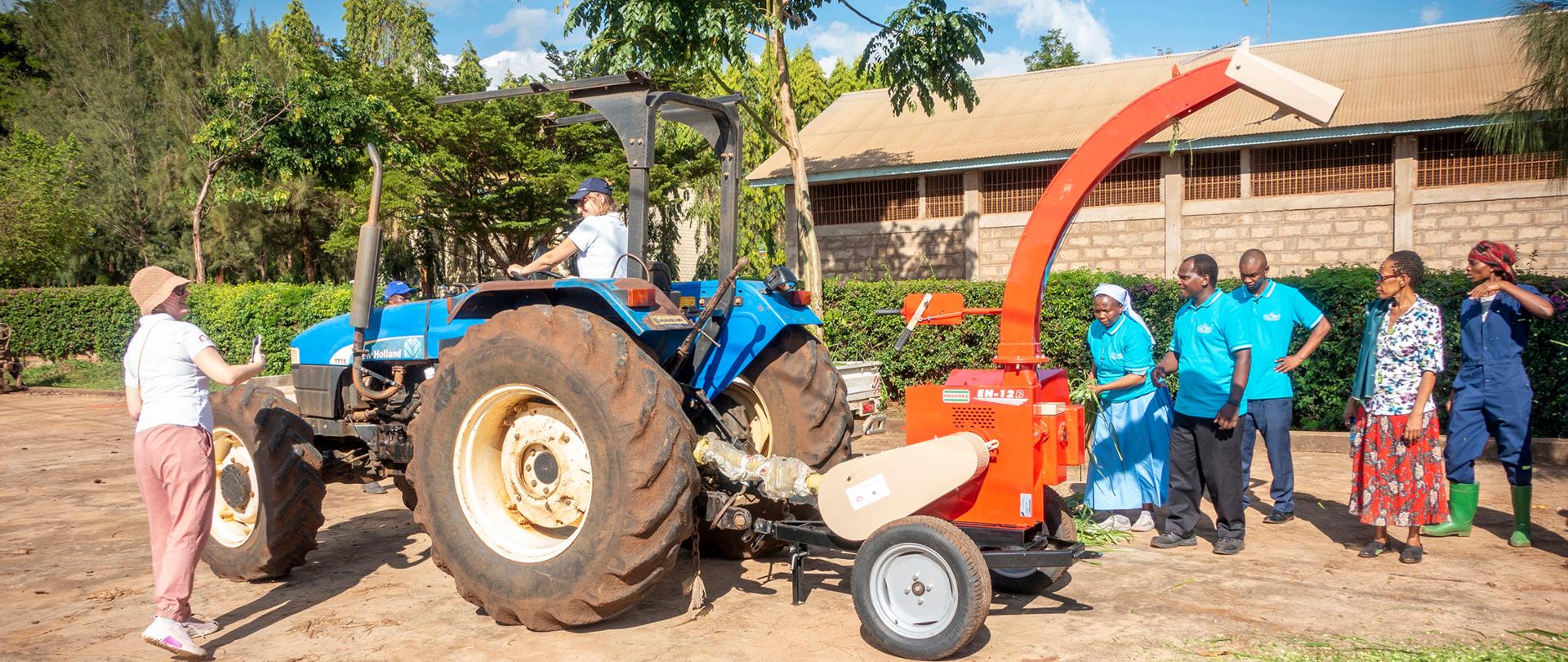 Kobieta prowadzi traktor, którego pracę obserwuje 5 osób, z boku stoi kobieta, która robi zdjęcie traktora