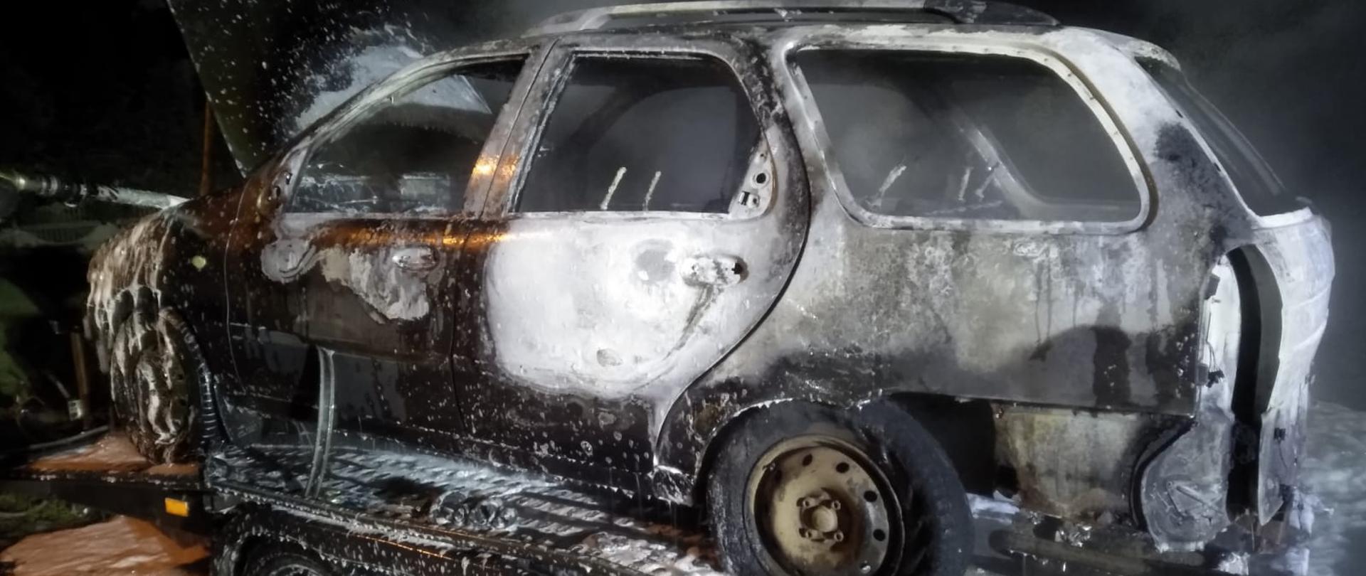 Kompletnie spalony samochód osobowy stojący na lawecie samochodowej pokryty pianą gaśniczą.