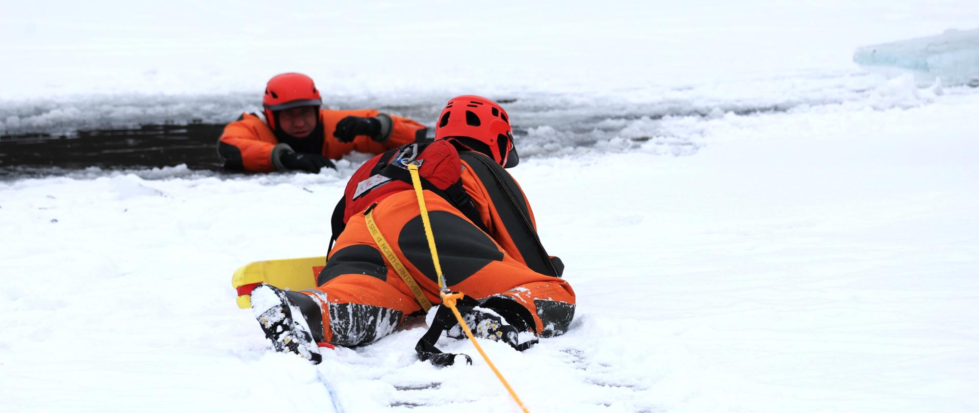 Szkolenie z zakresu ratownictwa lodowego - strażak w wodzie w przeręblu, a drugi czołga się do niego aby ćwiczyć jego ratowanie