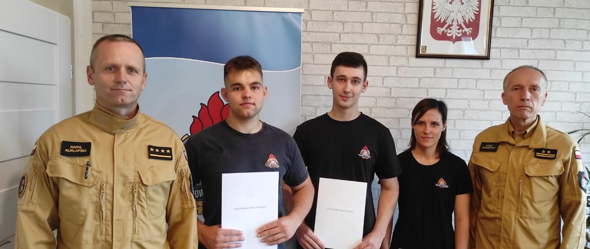 Zdjęcie przedstawia 5 strażaków w biurze komendanta, podczas wręczania aktu mianowania na strażaka. strażacy trzymają w ręku dokumenty