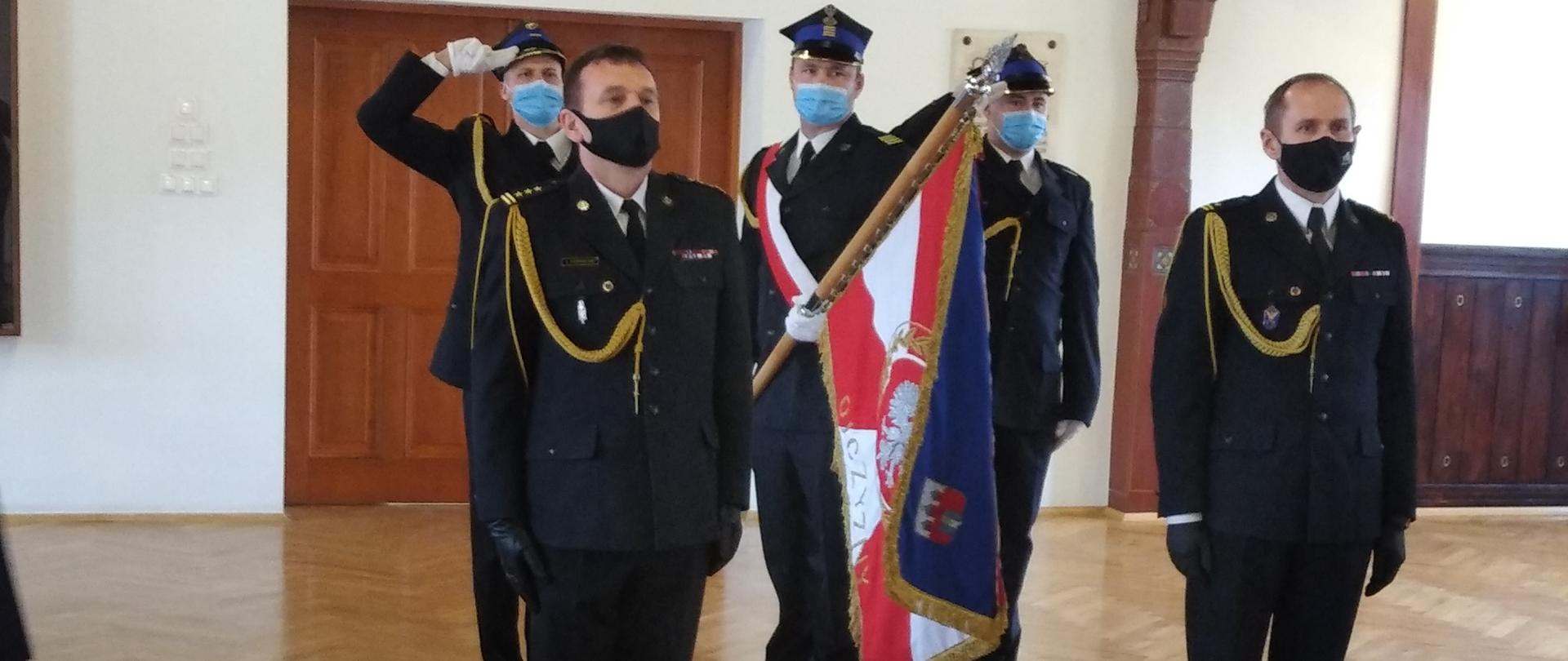 Na pierwszym planie od lewej strony: st. bryg. Jacek Podwolski- zdający obowiązki Zastępcy Komendanta Miejskiego PSP w Bydgoszczy, mł. bryg. Tomasz Czajka- przejmujący obowiązki Zastępcy Komendanta Miejskiego PSP w Bydgoszczy, obydwoje w mundurach wyjściowych i czarnych maseczkach na twarzy. W tle poczet sztandarowy KM PSP w Bydgoszczy, poczet składa się z 3 funkcjonariuszy PSP ubranych w mundury wyjściowe, środkowa osoba z pocztu pochyla sztandar w kierunku przejmującego i zdającego obowiązki. Całość odbywa się w pomieszczeniu świetlicy JRG1 KM PSP w Bydgoszczy. 