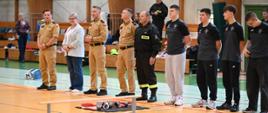 Szereg przedstawicieli straży pożarnej w beżowych i czarnych mundurach oraz gości w strojach wizytowych i sportowych stojących podczas zbiórki na parkiecie hali sportowej.