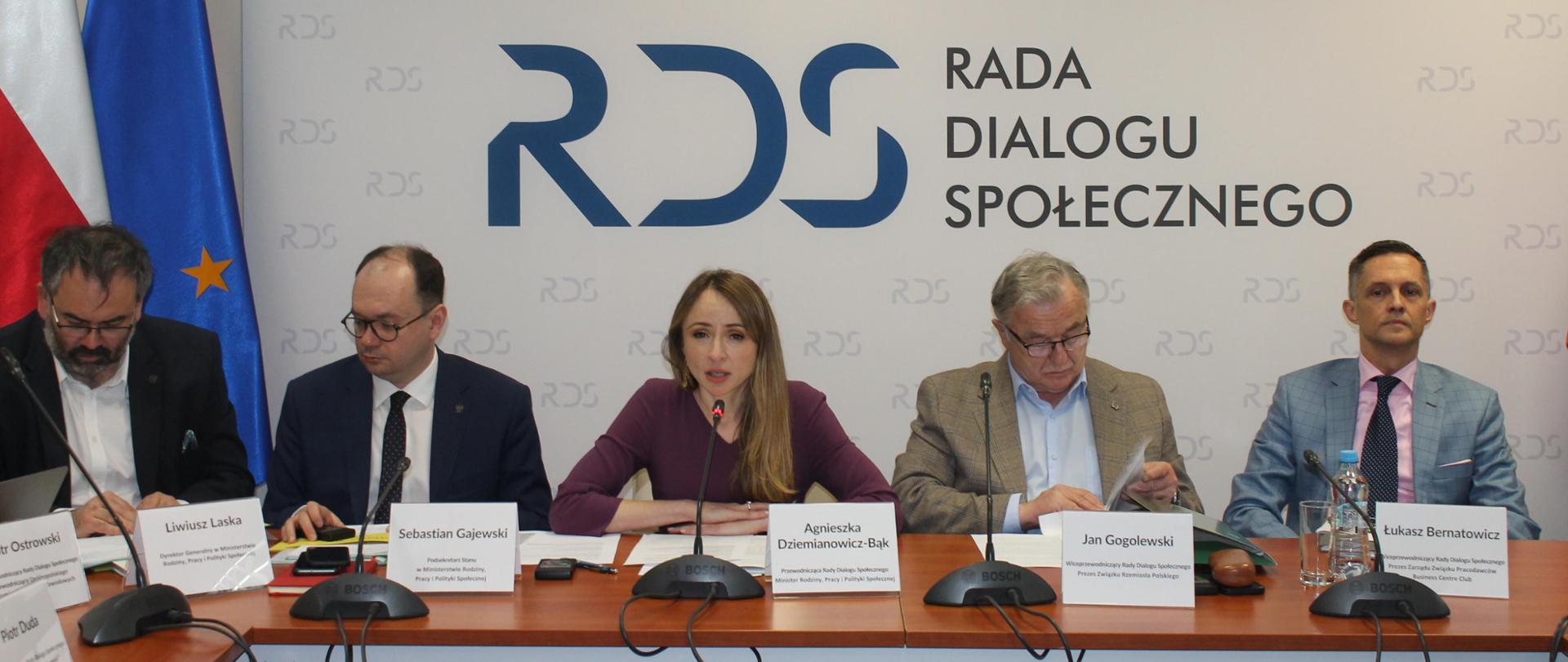 Na zdjęciu widnieje stół prezydialny za którym siedzą od lewej strony: Liwiusz Laska – Dyrektor generalny w MRPiPS, Sebastian Gajewski – podsekretarz Stanu w MRPiPS, Agnieszka Dziemianowicz-Bąk Ministra Rodziny, Pracy i Polityki Społecznej, Przewodnicząca Rady Dialogu Społecznego, Jan Gogolewski – Prezes Związku rzemiosła Polskiego, Łukasz Bernatowicz – Prezes Zarządu BCC. W tle siedzących znajduje się baner z napisem Rada Dialogu Społecznego. Po lewej stronie znajdują się flagi Polski i UE. Nad banerem godło Polski.