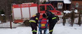 Na zdjęciu widać strażaków OSP w ubraniach koszarowych którzy pchają wózek do samochodu ratowniczego. W oddali widać dom. Aura jest zimowa.