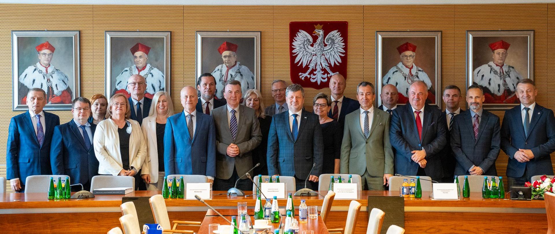 Zdjęcie zbiorowe, pod ścianą na której wisi rząd portretów stoi grupa kilkunastu osób, pośrodku minister Wieczorek.