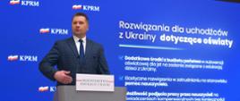 Minister Czarnek przemawia do mikrofonu na tle niebieskiej ściany z napisem Rozwiązania dla uchodźców z Ukrainy dotyczące oświaty.