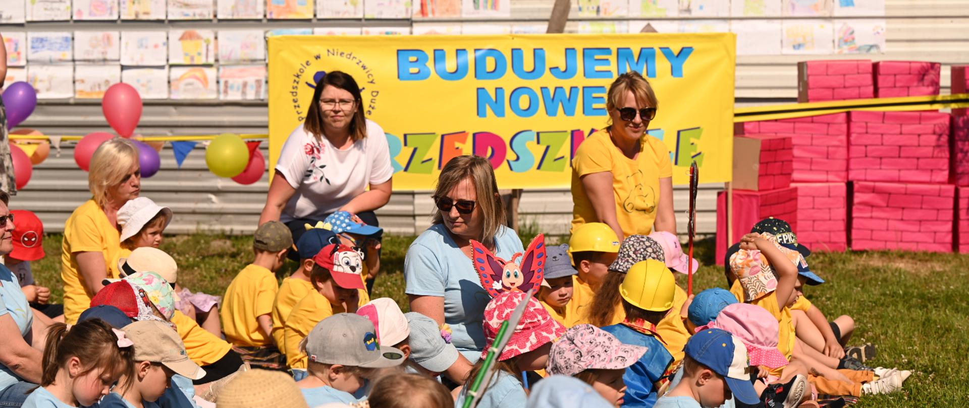 Kolorowo ubrane dzieci siedzą na trawniku, za nimi napis Budujemy nowe przedszkole.