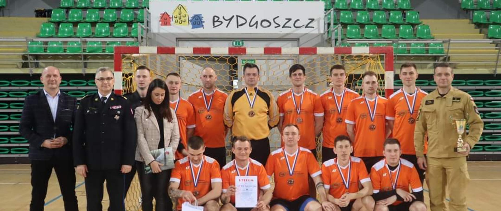 Zdjęcie przedstawia strażaków z KP PSP Radziejów w strojach sportowych na tle trybuny hali sportowej. Strażacy mają na szyjach medale za zajęcie trzeciego miejsca w rozgrywkach. Po lewej stronie w umundurowaniu wyjściowym stoi kujawsko-pomorski Komendant Wojewódzki PSP, po prawej stronie w umundurowaniu służbowym z pucharem w ręku stoi Komendant Powiatowy PSP z Radziejowa.