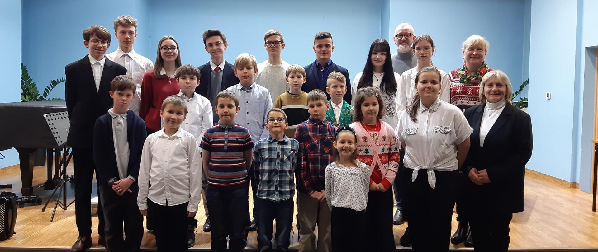 Na zdjęciu widać grupę osób pozujących wspólnie do zdjęcia w pomieszczeniu o niebieskich ścianach. W tle znajduje się scena w sali kameralnej oraz akordeony, fortepian i organy.
Grupa składa się z uczniów i nauczycieli. Wszyscy są elegancko ubrani, niektórzy w koszule, inni w swetry lub bluzki. Osoby stoją w kilku rzędach: dzieci z przodu, starsi uczniowie i dorośli z tyłu.