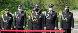 Zdjęcie przedstawia 5 strażaków OSP stojących w szeregu w galowych mundurach, a przed nimi stoi 1 strażak PSP - Komendant Wojewódzki PSP w Łodzi. Panowie stoją na placu z kostki betonowej, za nimi widać zielone drzewa, a przed nimi znajduje się czerwona taśma odgradzająca. 
