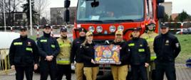 Strażacy na tle samochodu strażackiego trzymają baner WOŚP z napisem gasimy ogień rozpalamy serca.