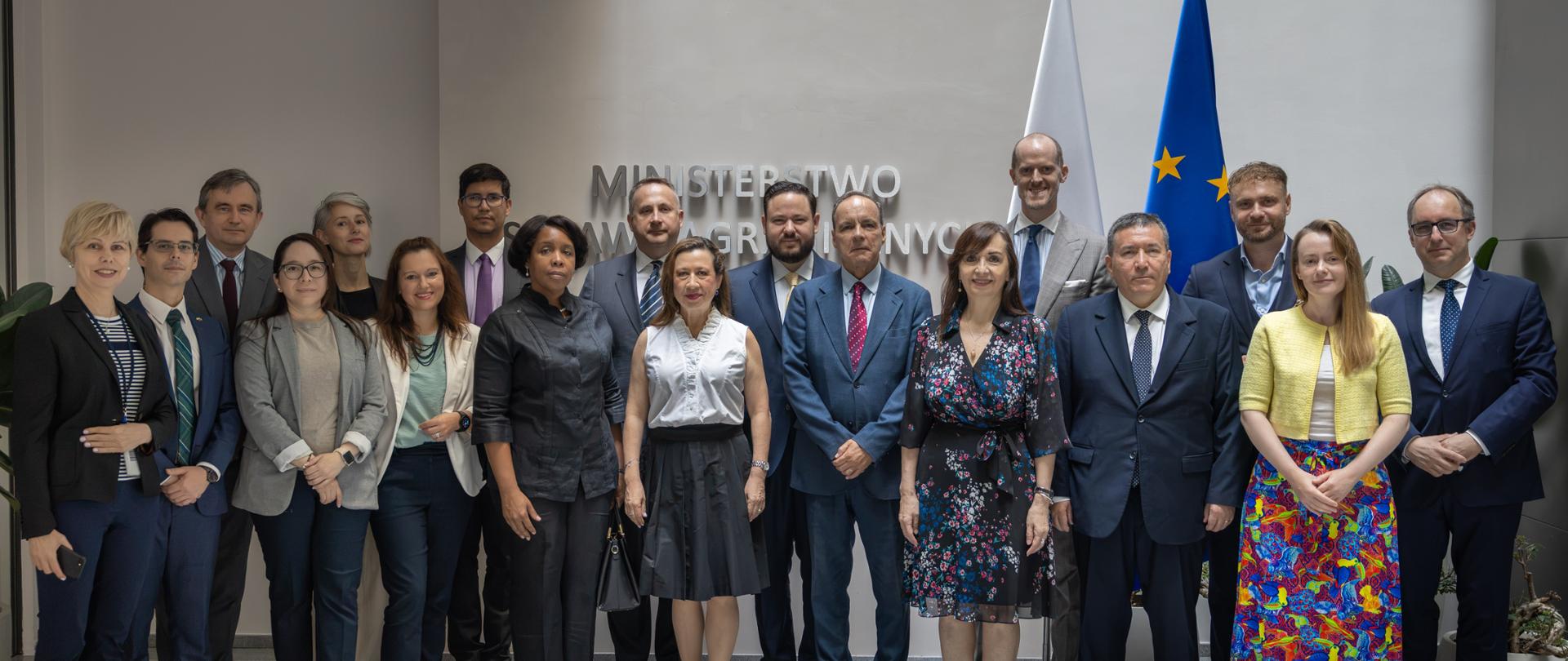 Family photo in the lobby of the Ministry of Foreign Affairs. People stand in two rows in front of a wall displaying the Ministry's name, with the flags of Poland and the European Union visible in the background. Everyone is facing the camera.