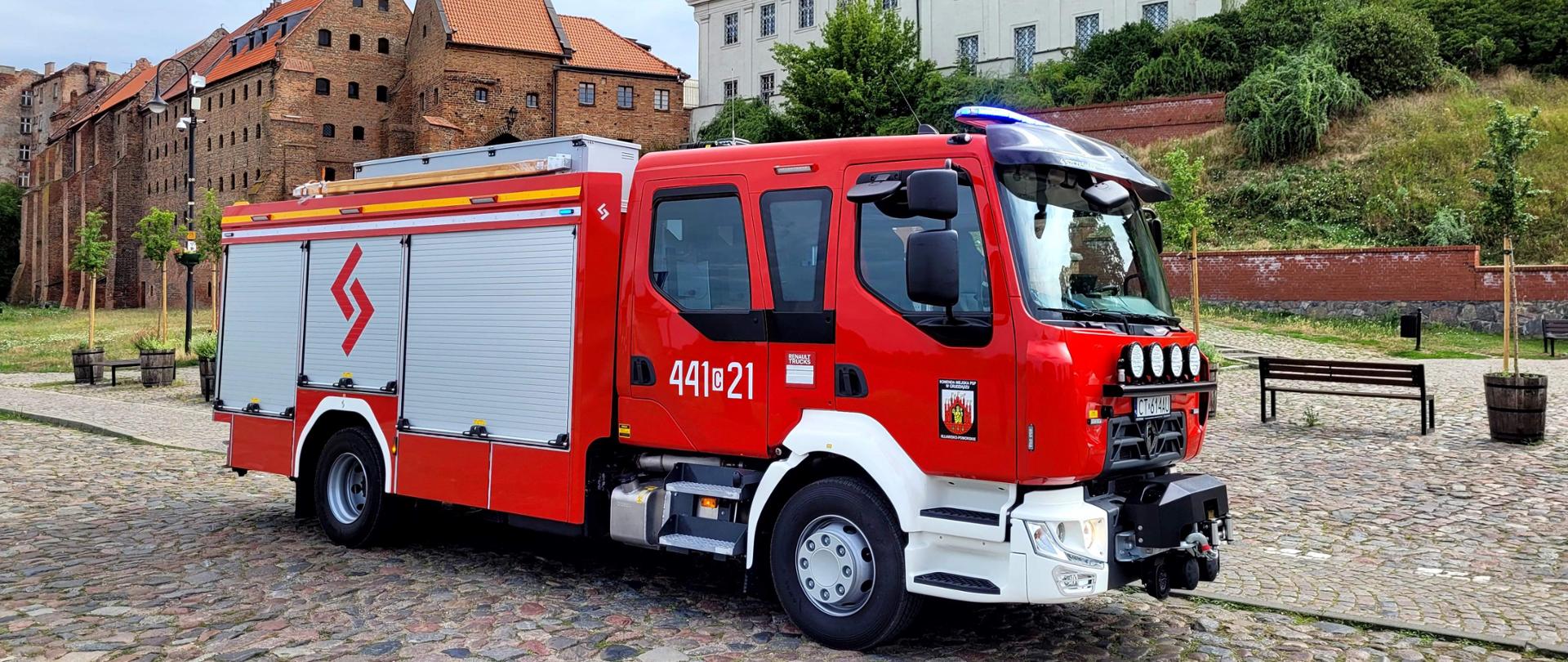 Czerwony ciężarowy samochód strażacki stojący na bruku przed zabytkowymi gotyckimi spichlerzami.