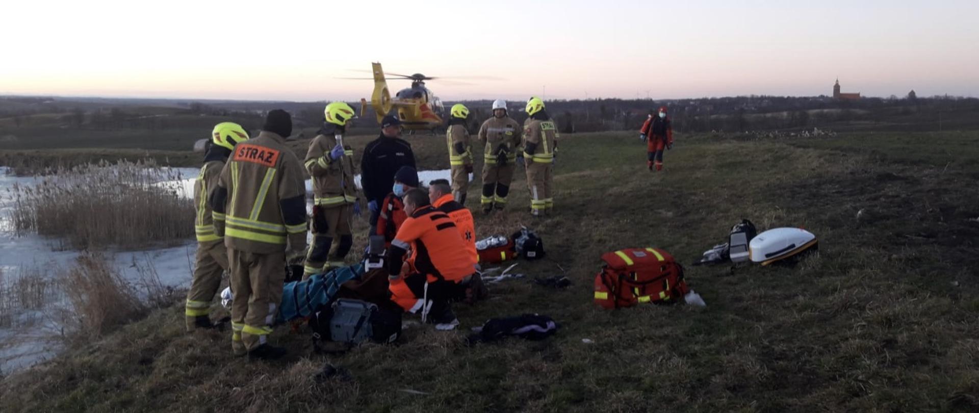 Zdjęcie przedstawia strażaków w ubraniach specjalnych koloru piaskowego, ratowników medycznych w ubraniach koloru czerwonego oraz jaskrawo-czerwonego oraz policjanta w niebieskim mundurze. Służby udzielają pomocy mężczyźnie wyciągniętemu z wody. W tle widać przydomowy staw oraz helikopter LPR koloru żółtego.