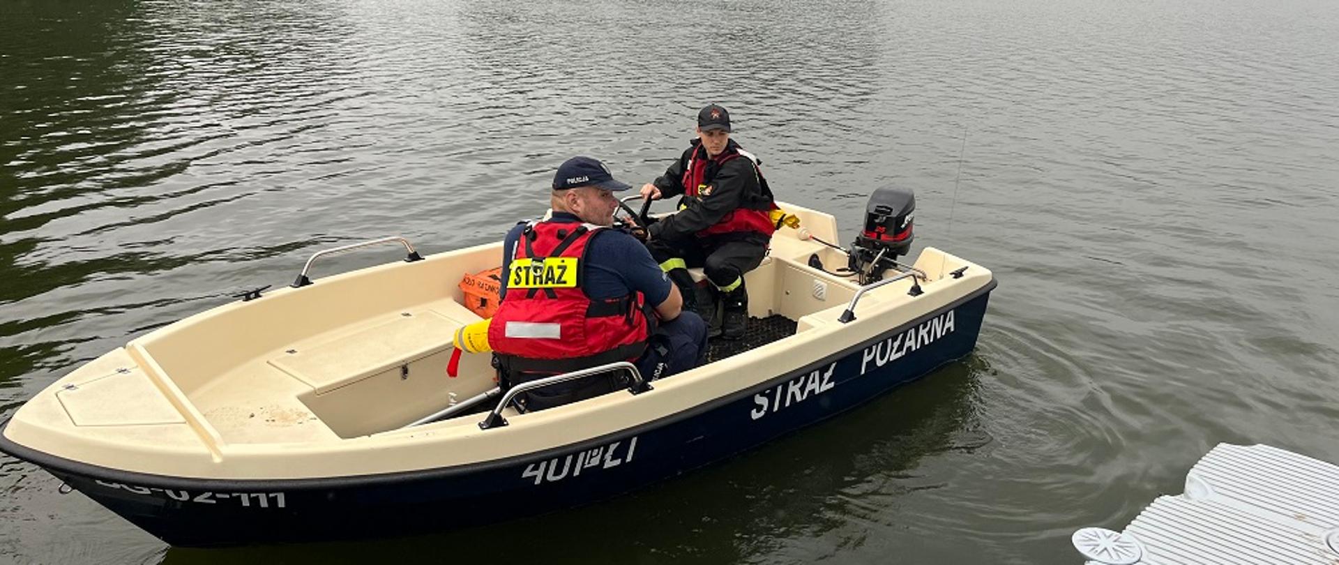 Zdjęcie przedstawia strażaków i policjantów patrolujących jezioro.
W tle jezioro i las.
