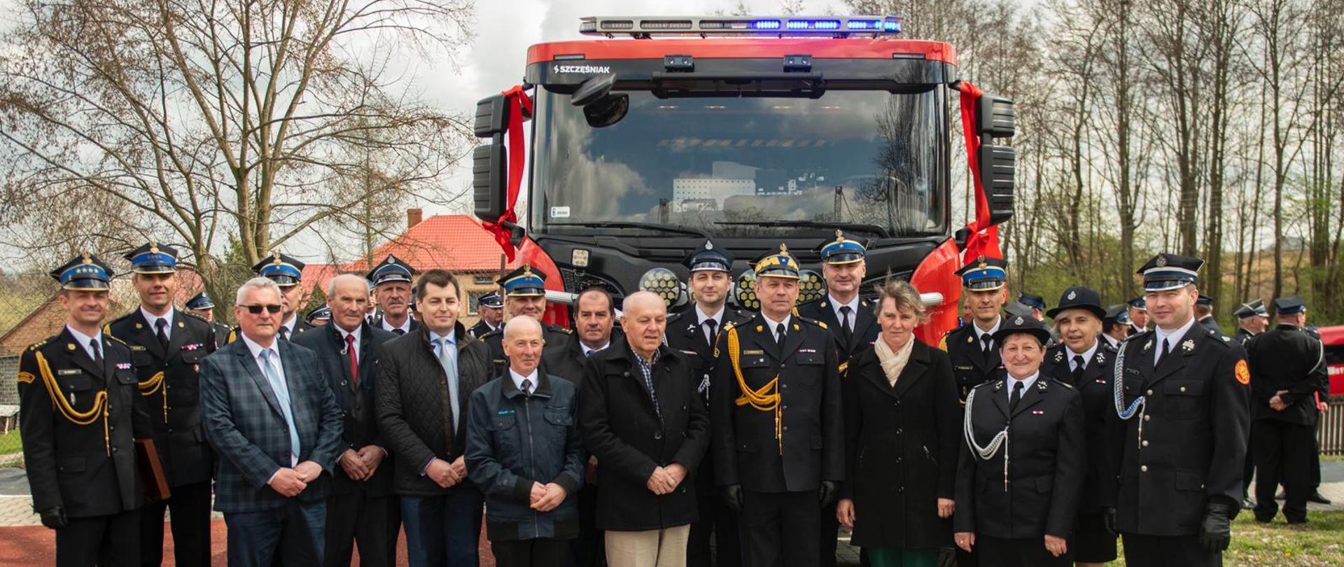 Grupa osób (strażacy i osoby cywilne) uczestnicy uroczystości pozują grupowo do zdjęcia pamiątkowego, za nimi stoi samochód ratowniczo - gaśniczy 