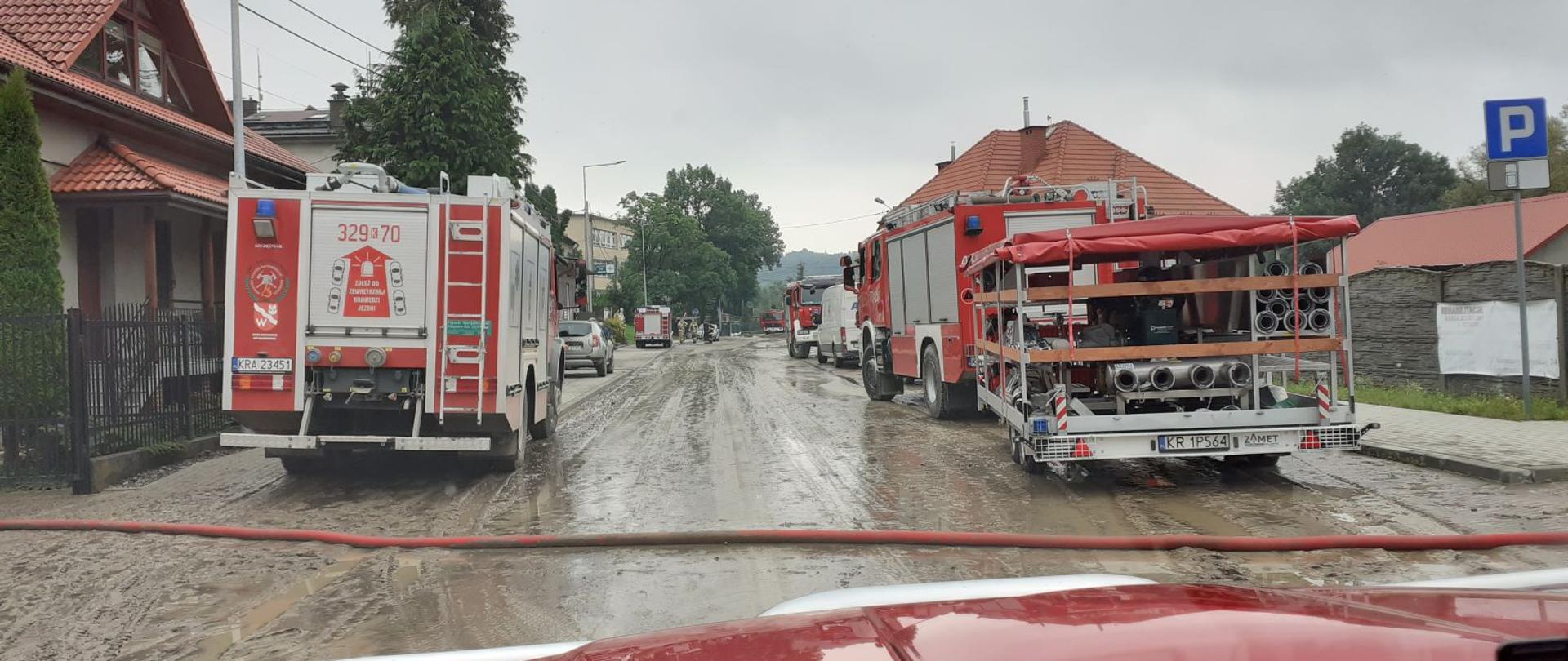 Zdjęcie przedstawia drogę gminną i centrum miejscowości Głogoczów po przejściu fali błota. Po obydwu stronach drogi stoją samochody strażackie, z których sprzęt wykorzystywany jest do wypompowywania wody z domów.