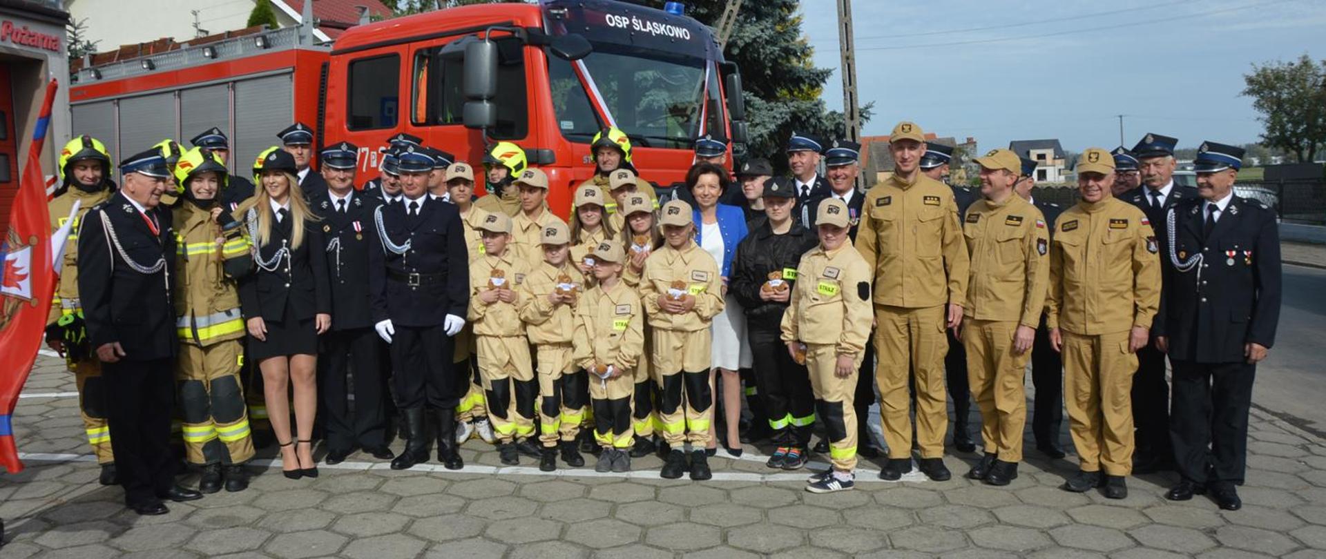 Zdjęcie przedstawia Komendantów PSP, strażaków z OSP oraz dzieci i młodzież z MDP na tle przekazanego nowego ciężkiego pojazdu pożarniczego.