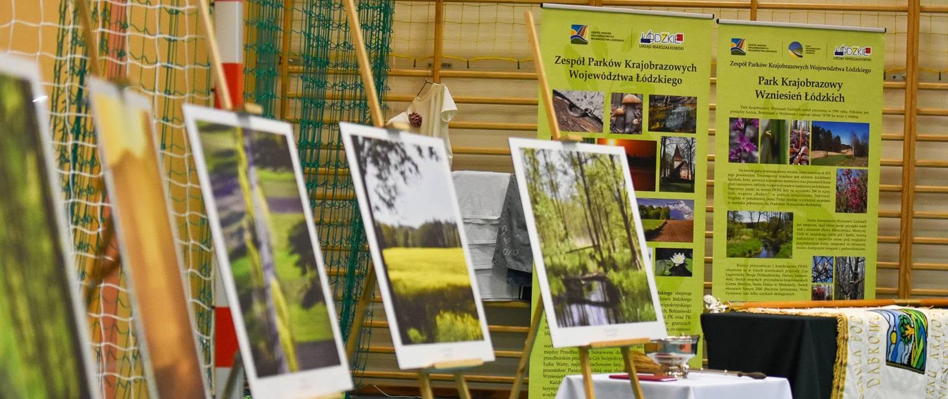 Ekspozycja fotografii przedstawiających przyrodę Parku Krajobrazowego Wzniesień Łódzkich ustawiona na sztalugach w sali gimnastycznej. W tle znajdują się plansze informacyjne o parku oraz stół przygotowany na uroczystość nadania szkole patrona
