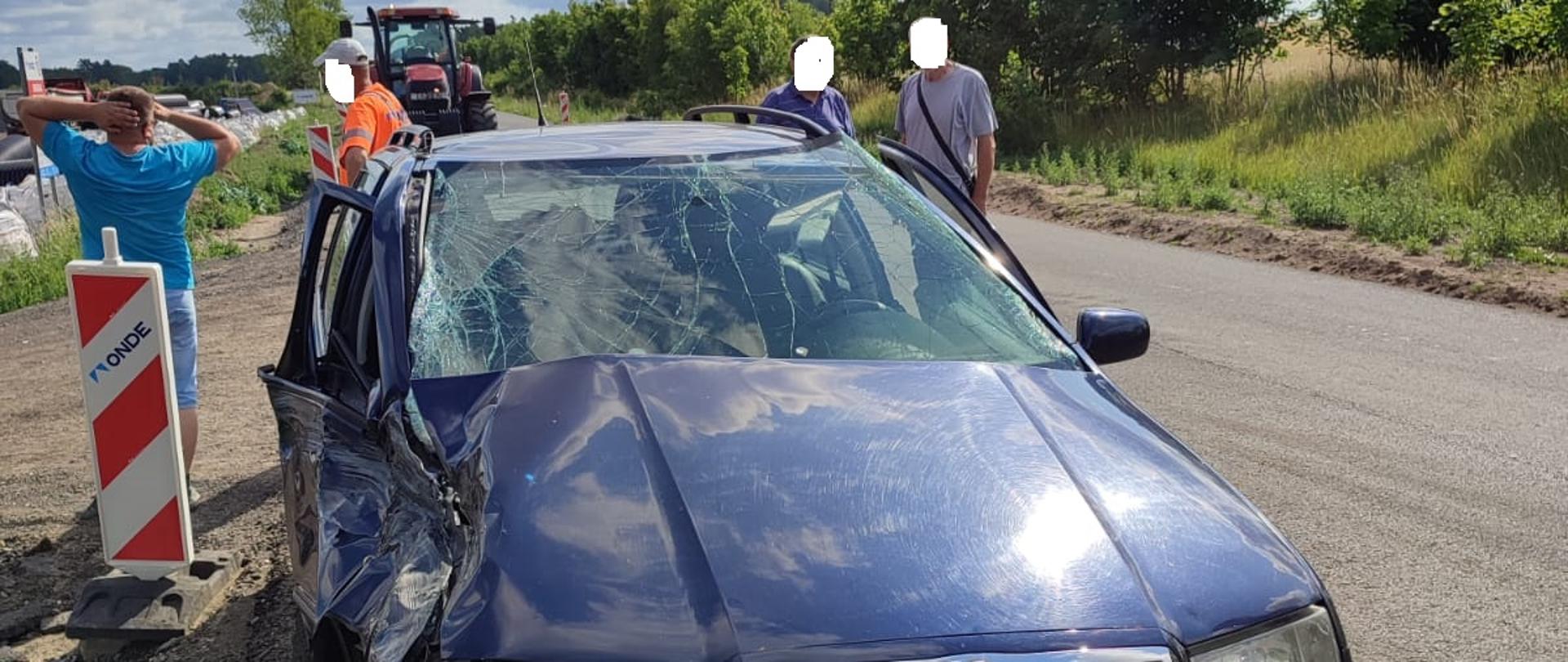 Zdjęcie przedstawia niebieski samochód osobowy z uszkodzonym lewym bokiem po kolizji z ciągnikiem rolniczym, który znajduje się za nim. Obok auta stoją ludzie.