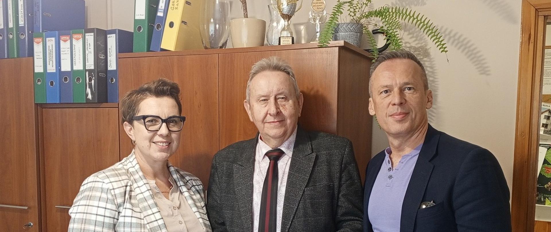 Na zdjęciu w biurze znajdują się trzy osoby: kobieta i dwóch mężczyzn, stojący za drewnianym biurkiem i przed regałem z segregatorami.Kobieta po lewej stronie ma na sobie biało-szary garnitur w kratę i okulary.Mężczyzna w środku, starszy, w szarej marynarce i krawacie, trzyma w rękach małe, kolorowe, podłużne pudełko.Mężczyzna po prawej stronie ubrany jest w granatowy blezer, jasnofioletową koszulę i dżinsy.W tle widoczne są rośliny doniczkowe oraz trofea na górze szafek. Na biurku, przed postaciami, leżą dokumenty i czerwony zszywacz.