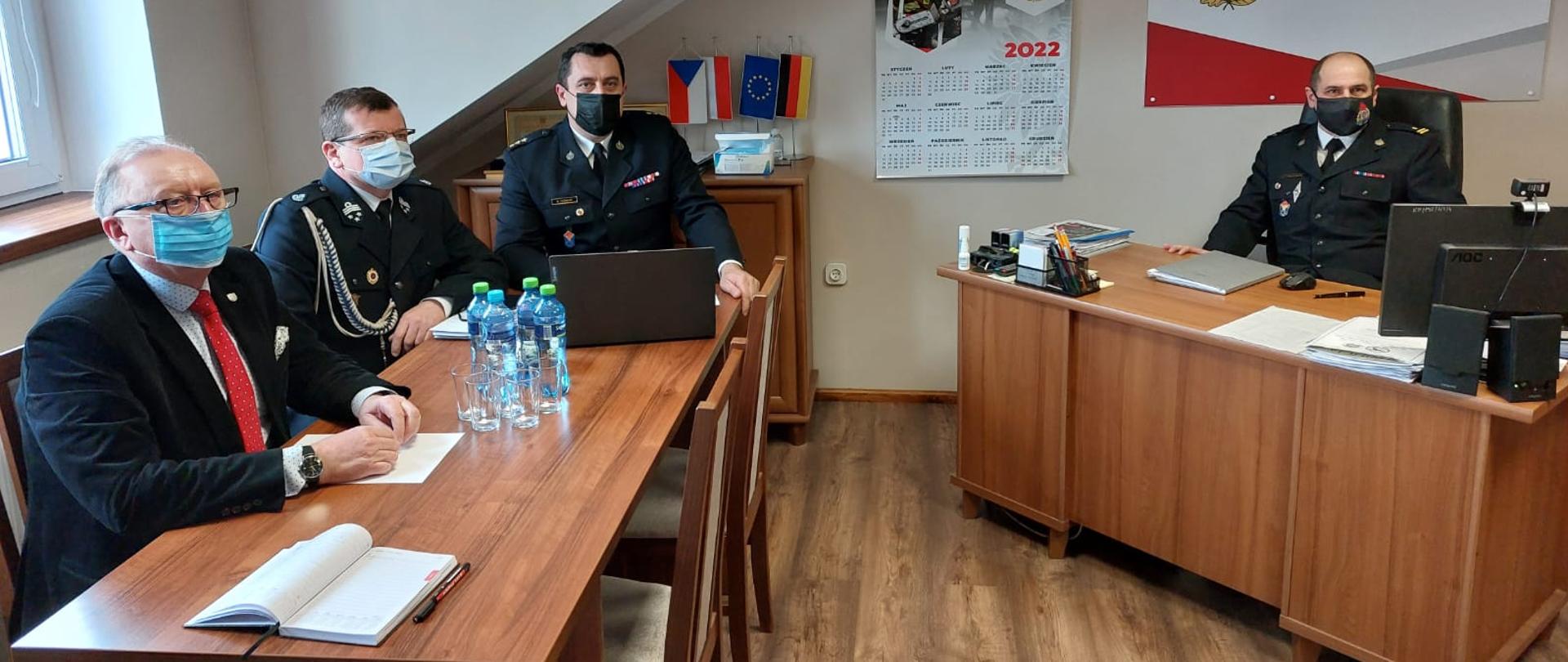 Pomieszczenie biurowe. Z prawej strony stoi biurko na którym znajduje się monitor komputerowy, kilka dokumentów, telefon bezprzewodowy. Za biurkiem siedzi Komendant Powiatowy PSP w Zgorzelcu ubrany w mundur wyjściowy. Na ścianie za biurkiem wisi baner z logiem PSP oraz danymi kontaktowymi KP PSP w Zgorzelcu. Z lewej strony stoi stół (na stole kilka butelek z wodą mineralną, notatniki oraz otwarty komputer przenośny), za którym siedzi wice starosta zgorzelecki w ubraniu cywilnym obok niego drug OSP w mundurze wyjściowym OSP oraz Z-ca Komendanta Powiatowego PSP w mundurze wyjściowym. Wszytki osoby na zdjęciu mają ubrane maseczki ochronne