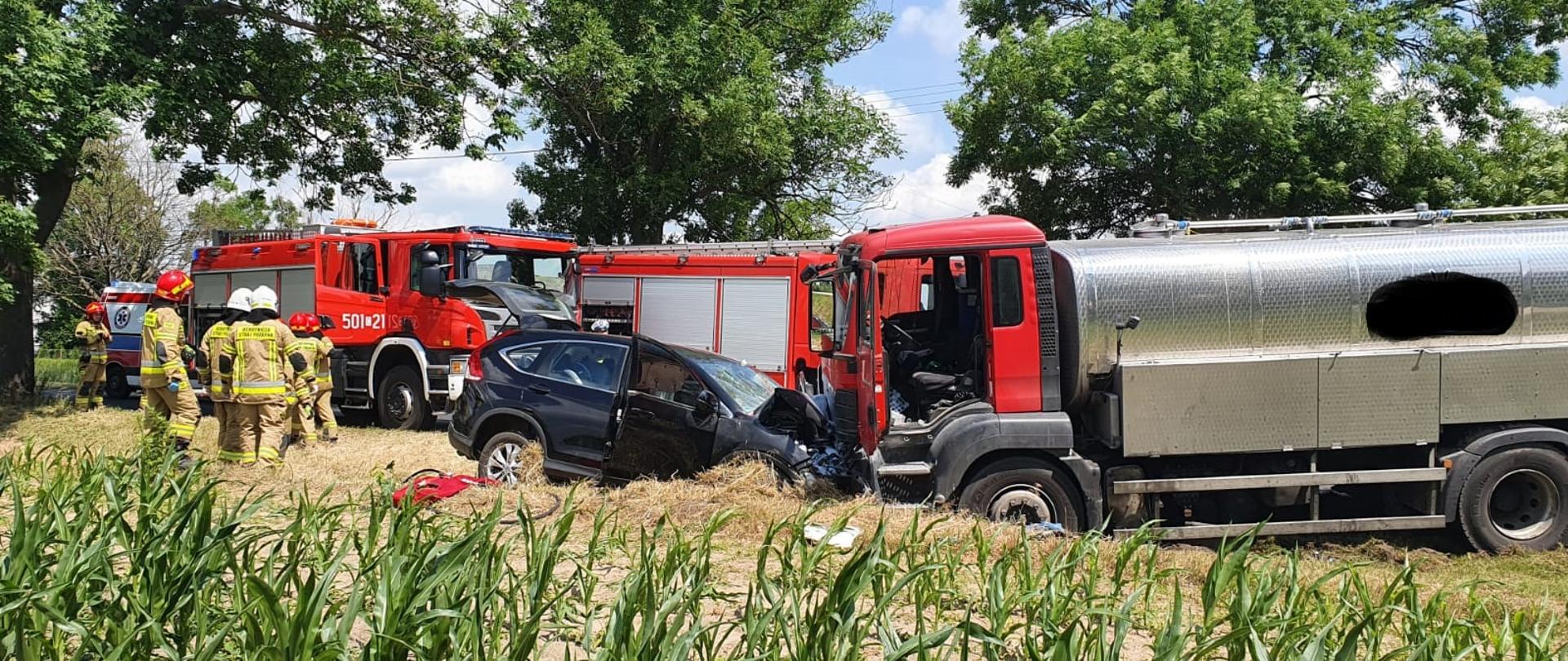 Zdjęcie przedstawia samochód osobowy marki Honda ( koloru ciemnego) i samochód ciężarowy marki MAN cysterna do przewozu mleka ( kabina koloru czerwonego, zbiornik srebrny) po wypadku drogowym. Na drugim planie dwa samochody pożarnicze (koloru czerwonego). Obok pojazdów stoją ubrani w piaskowe mundury bojowe strażacy. Na głowach mają założone białe i czerwone hełmy