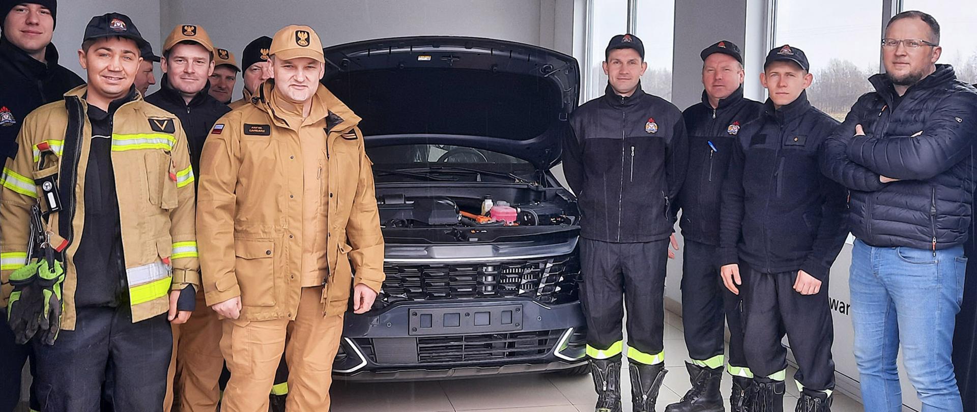 Na pierwszym planie grupa strażaków znajdujących się w garażu pozuje do zdjęcia, między nimi stoi samochód z otwartą maską. Prawa strona oszklona jest w dwa duże przejrzyste okna. 