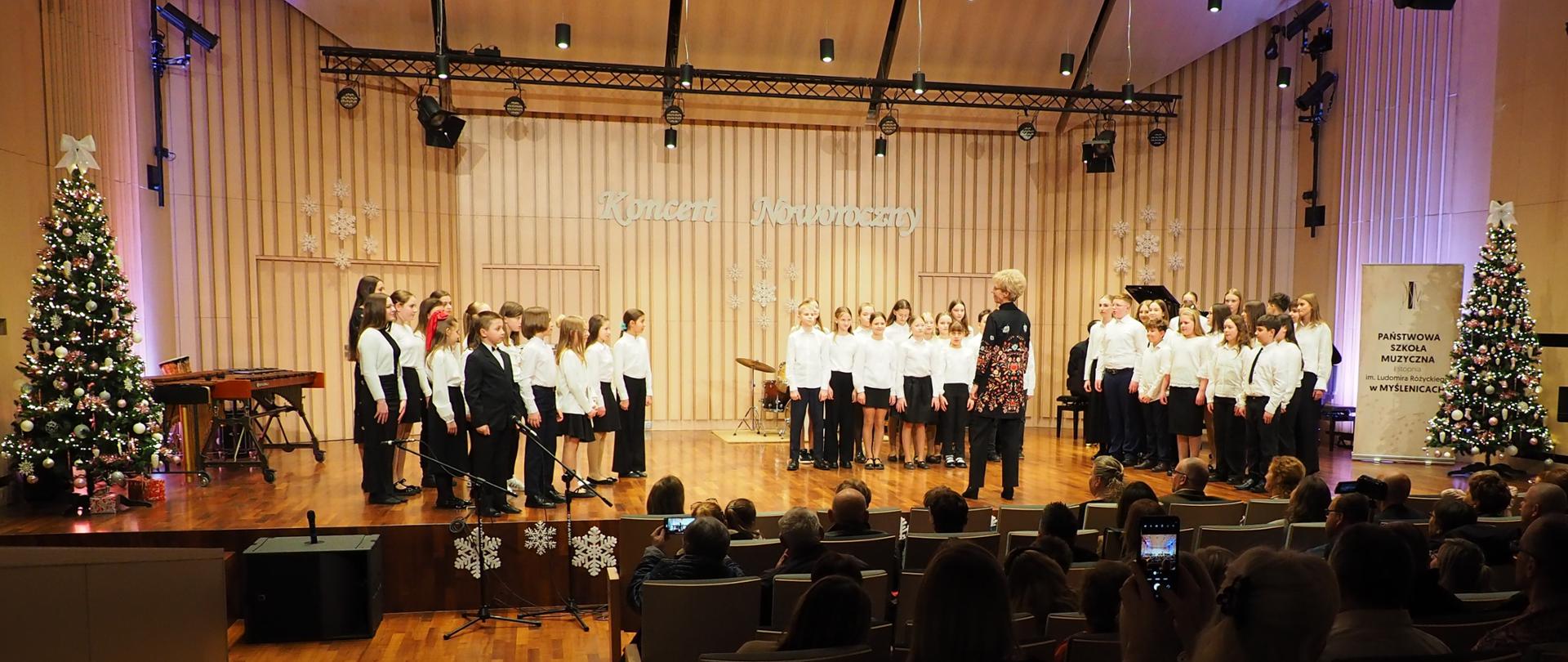 Chór szkolny na scenie sali koncertowej z dyrygentem, w tle napis Koncert Noworoczny, z prawej strony zdjęcia plakat z logo oraz napisem Państwowa Szkoła Muzyczna I stopnia im. Ludomira Różyckiego w Myślenicach, po lewej stronie instrumenty perkusyjne, po bokach sceny choinki świąteczne, na pierwszym planie siedząca publiczność.