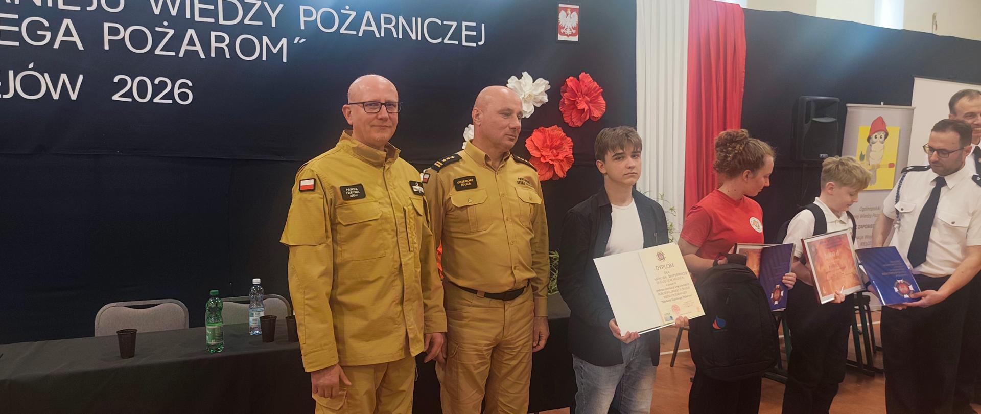 Na pierwszym planie uczestnicy turnieju, w rękach trzymają dyplomy, w centralnej części fotografii dwóch funkcjonariuszy PSP w żółtych ubraniach służbowych
