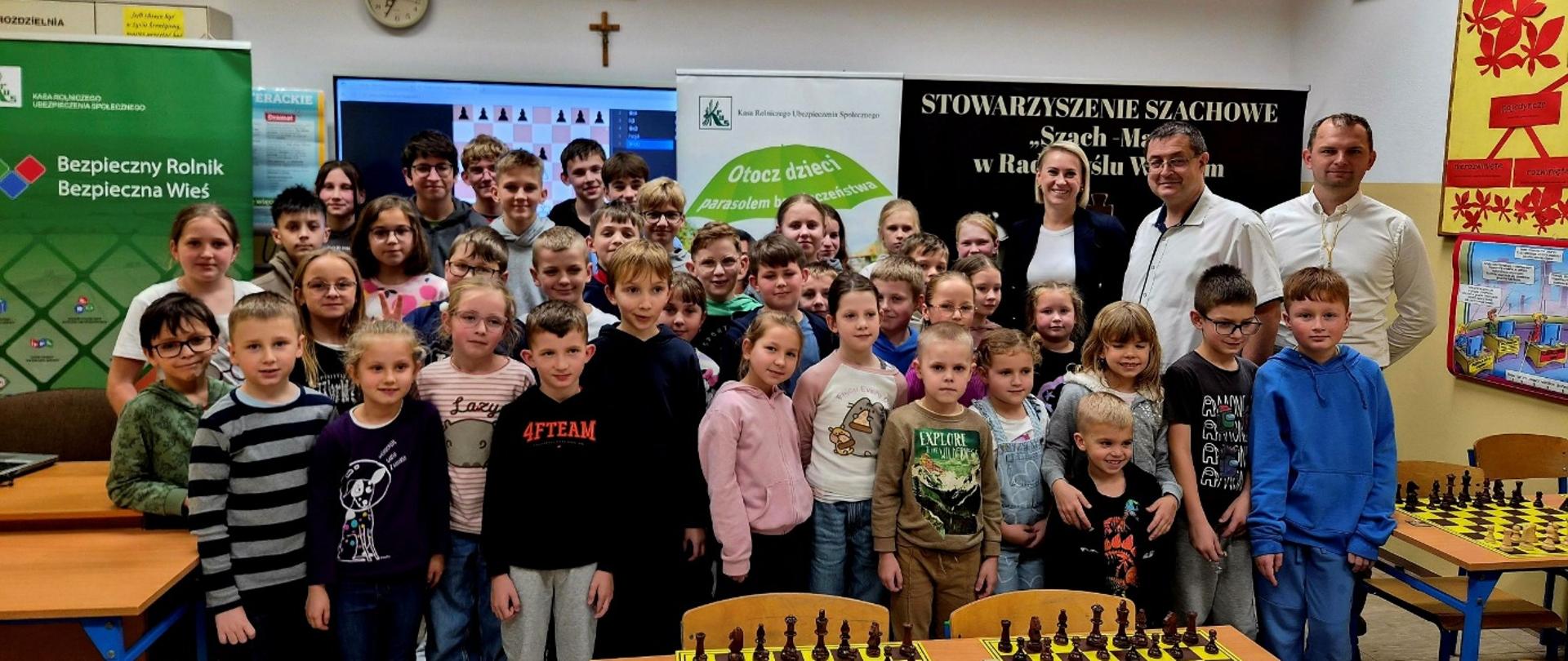 Grupa osób pozuje do zdjęcia – są to: Uczestnicy turnieju, pracownicy PT KRUS w Mielcu: Aneta Cebula, Robert Żyła i sędzia główny Pan Przemysław Kopacz. Za nimi znajdują się rollupy KRUS oraz Stowarzyszenia Szachowego „Szach-Mat” w Radomyślu Wielkim.