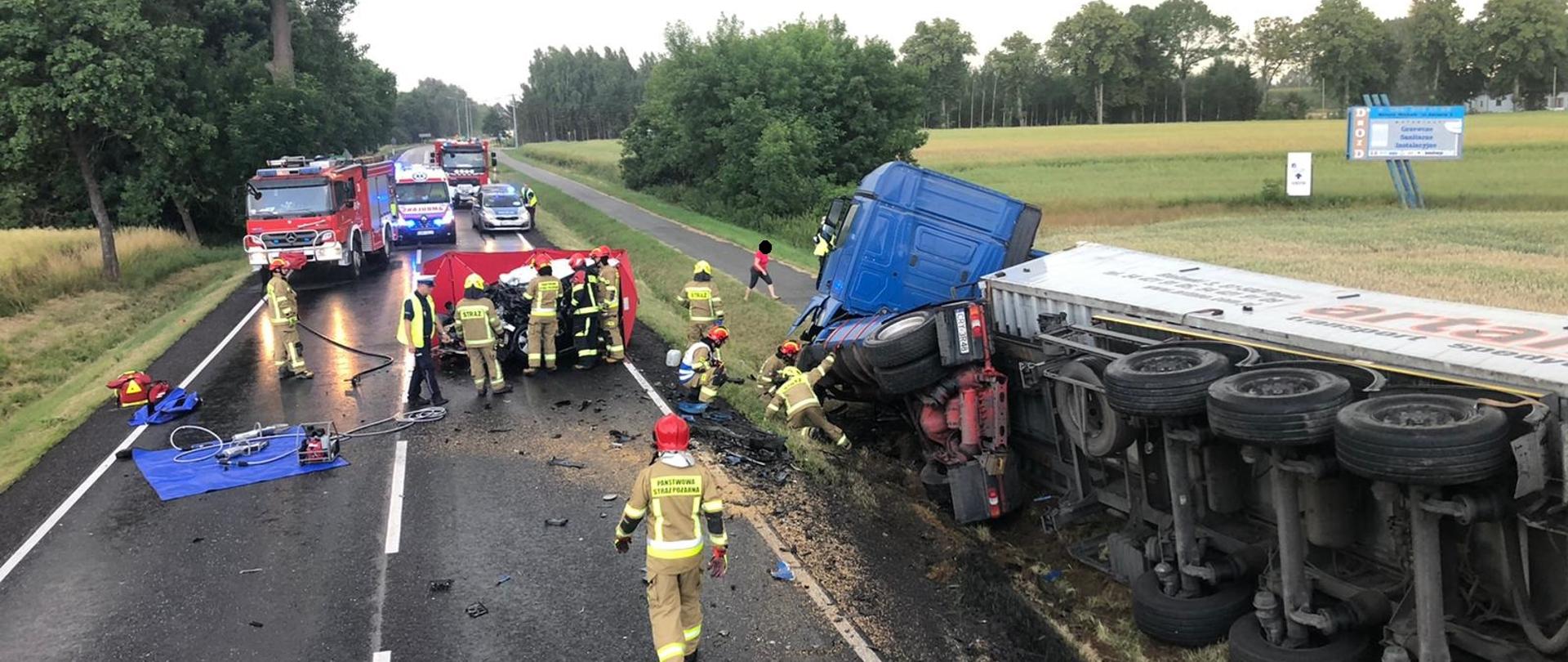 Fotografia przedstawia służby ratownicze podczas działań przy wypadku drogowym. Na fotografii widać rozbite wraki pojazdów biorących udział w zdarzeniu oraz pojazdy straży pożarnej, policji oraz pogotowia ratunkowego.