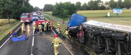 Fotografia przedstawia służby ratownicze podczas działań przy wypadku drogowym. Na fotografii widać rozbite wraki pojazdów biorących udział w zdarzeniu oraz pojazdy straży pożarnej, policji oraz pogotowia ratunkowego.