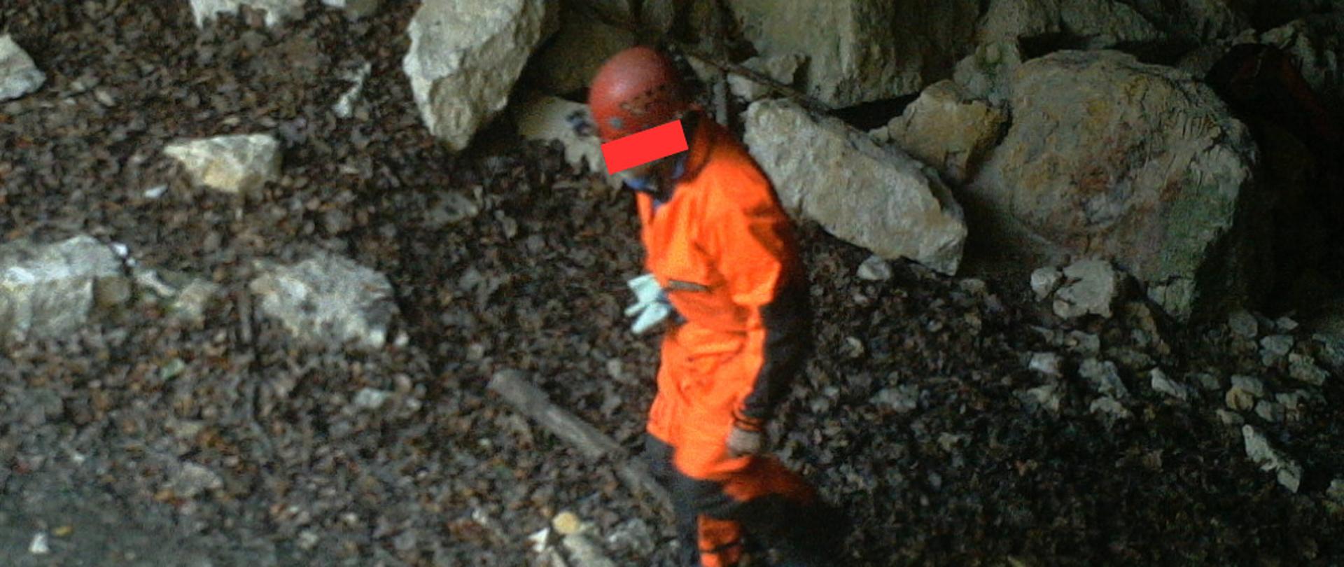 Osoba z kaskiem i w specjalnym ubraniu wskazującym na grotołaza idąca przez jaskinię 