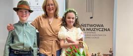 Zdjecie przedstawia uczennice w sukience trzymajaca w ręku kosz z tulipanami , ucznia ubranego w koszule koloru zielonego oraz granatowe spodnie na głowie ma kapelusz oraz nauczycielke ubrana w niebieskie spodnie, brązowa bluzke . W tle widac plakat z napisem Państwowa Szkoła Muzyczna w Zielonej Górze