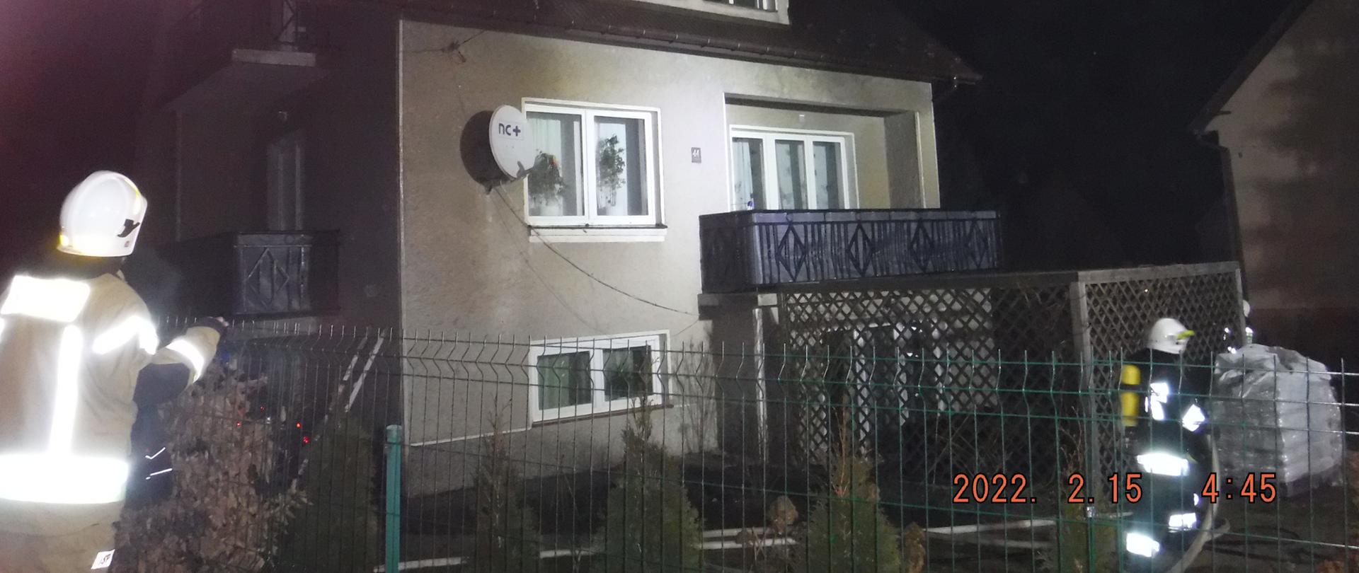 Zdjęcie przedstawia murowany dom w którym miał miejsce pożar. Z dachy budynku widać unoszący się biały dym. Po lewej stronie przy zielonym ogrodzeniu widać zwróconego w kierunku budynku strażaka ubranego w piaskowe ubranie specjalne i biały hełm. Po prawej stronie stoi strażak ubrany w czarne ubranie specjalne i biały hełm na plecach ma założoną aparat ochrony dróg oddechowych i w ręku trzyma wąż pożarniczy. Za strażakiem stoi paleta z workami. Na ścianie budynku i oknach widać odbicie światła pochodzące z sprzętu oświetleniowego zamontowanego na samochodach pożarniczych. po lewej stronie znajduje się przystawiona do budynku drabina.