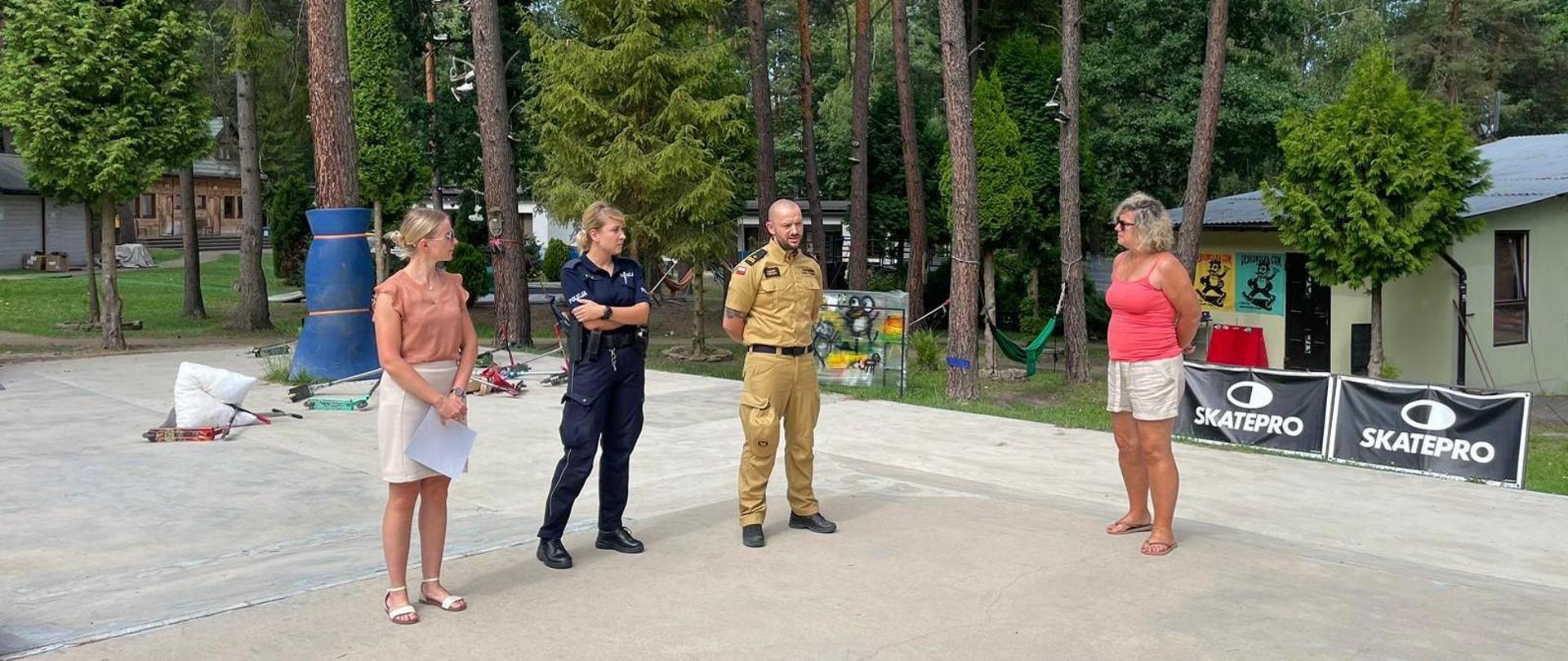 Na zdjęciu widać czworo dorosłych stojących na utwardzonym terenie pośród drzew, na świeżym powietrzu, prawdopodobnie w ośrodku rekreacyjnym lub obozowisku. Od lewej do prawej:
Kobieta o blond włosach, ubrana w jasną bluzkę i spódnicę, trzyma jakieś papiery.
Kobieta w ciemnoniebieskim mundurze, policjantka, z widoczną odznaką na piersi.
Mężczyzna w musztardowym ubraniu służbowym, oficer Państwowej Straży Pożarnej, z widocznym stopniem na pagonie koszuli.
Kobieta o blond włosach, ubrana w różową lub koralową bluzkę i jasne szorty.
W tle widać liczne wysokie sosny i inne zielone drzewa. Pomiędzy drzewami widać kilka małych drewnianych budynków. Po prawej stronie znajdują się dwa czarne banery lub znaki z białym napisem „SKATEPRO”. Oświetlenie sugeruje, że jest dzień.