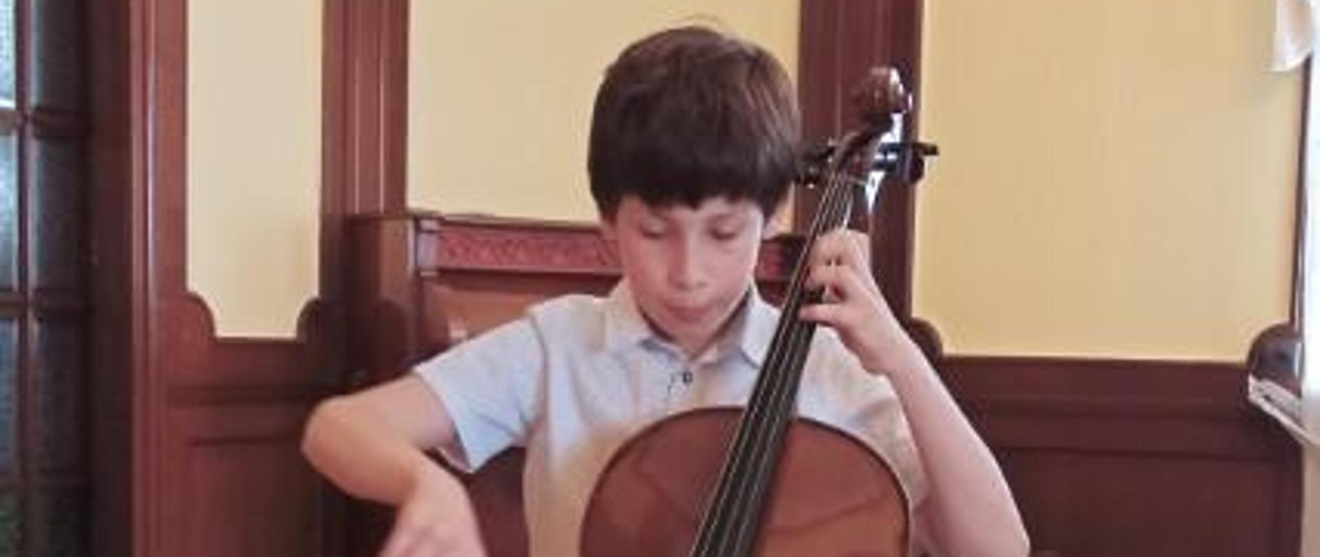 Zdjęcie kolorowe. Ciemnowłosy chłopiec w białej koszuli z nadrukiem w kropki gra na wiolonczeli. W tle ściana z ciemnobrązową boazerią.