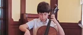 Zdjęcie kolorowe. Ciemnowłosy chłopiec w białej koszuli z nadrukiem w kropki gra na wiolonczeli. W tle ściana z ciemnobrązową boazerią.