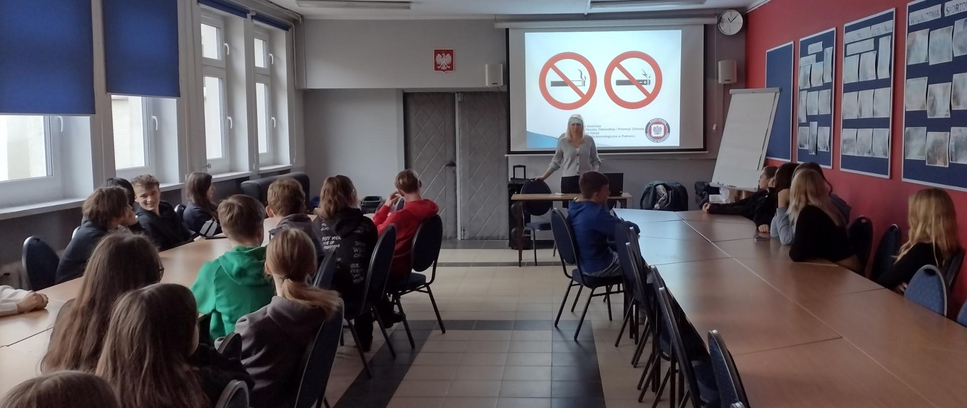 W dużej sali uczniowie siedzący przy stołach słuchają wykładu, w tle prezentacja wyświetlona na ekranie.