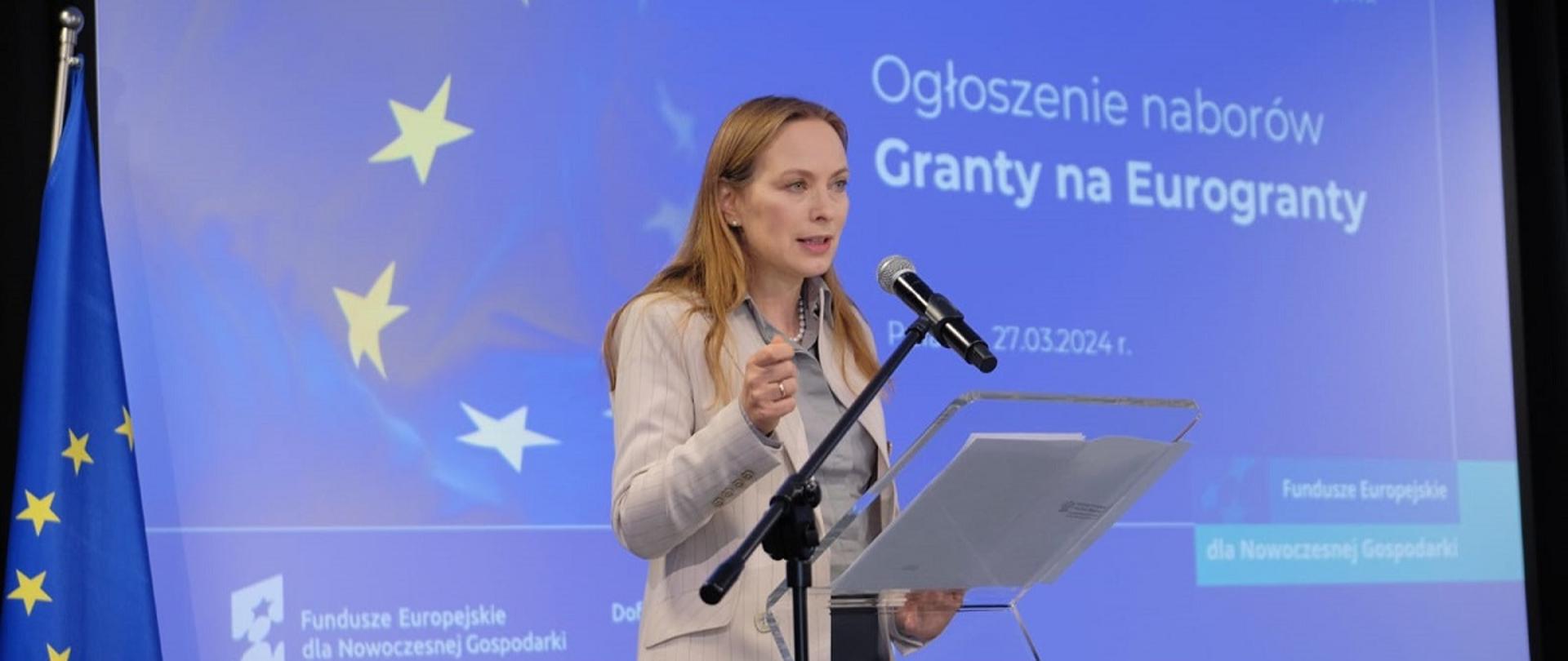 Minister funduszy i polityki regionalnej Katarzyna Pełczyńska-Nałęcz stoi przy pulpicie z mikrofonem, za jej plecami grafika z napisem informującym o naborach na Granty dla Eurograntów
