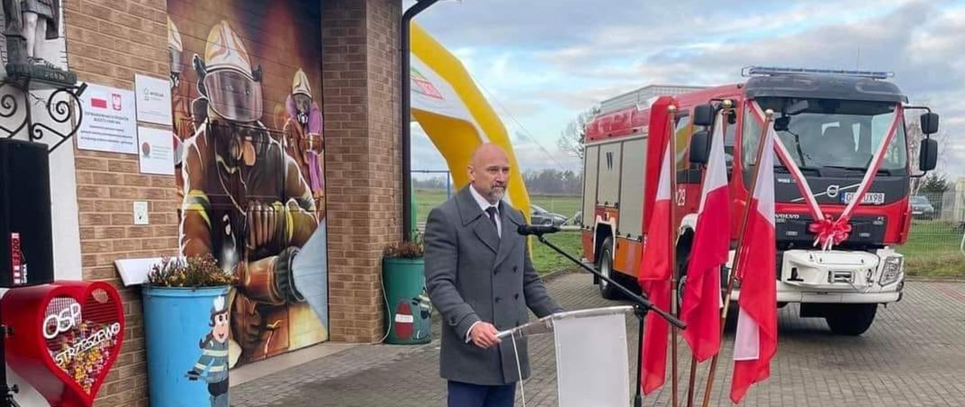 Zdjęcie przedstawia Wójta Gminy Wicko przemawiającego z mównicy na tle remizy OSP oraz przekazywanego samochodu ratowniczo-gaśniczego.