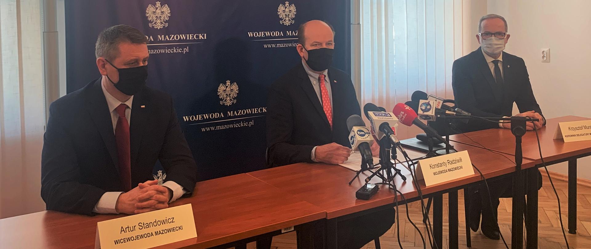 Wojewoda Mazowiecki, Wicewojewoda oraz kierownik delegatury Mazowieckiego Urzędu Wojewódzkiego w Radomiu siedzą przy stole prezydialnym podczas konferencji w budynku. Za nimi ciemnogranatowa ścianka z białym orłem i napisem Wojewoda Mazowieckie. Wojewoda Radziwiłł przemawia do ustawionych przed nim mikrofonów. 