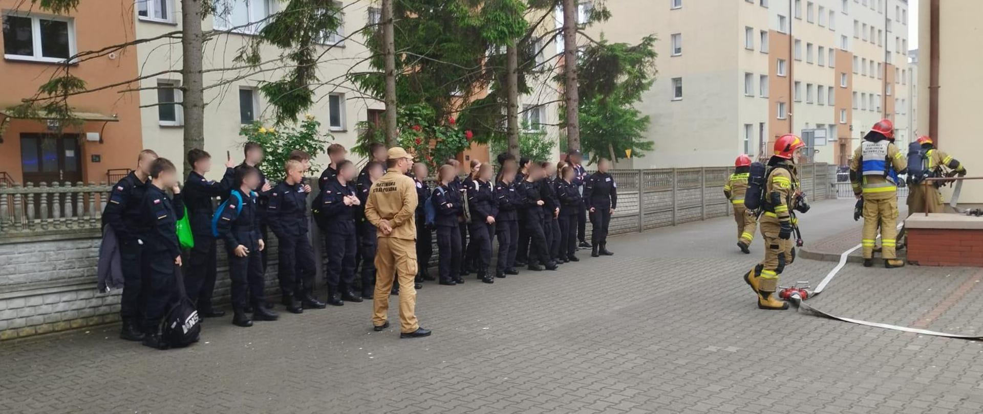Na zdjęciu widoczna klasa mundurowa w dwuszeregu. Przed frontem szyku stoi strażak w umundurowaniu służbowym koloru piaskowego. Od prawej strony widać pokaz akcji ratowniczo-gaśniczej. Strażacy wyposażeni w sprzęt ochrony dróg oddechowych, hełmy oraz ubrania specjalne koloru piaskowego prowadzą rozwinięcie do piwnicy budynku. Wszyscy stoją na kostce brukowej koloru szarego. W tle drzewa oraz bloki mieszkalne.