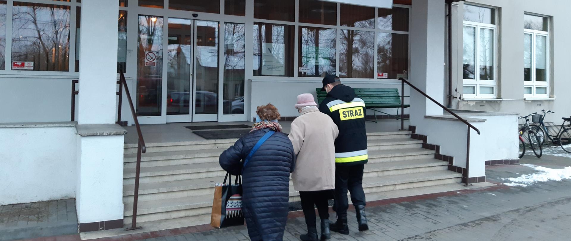 Na zdjęciu dwie kobiety oraz strażak, który jedną z nich trzyma pod rękę. Cała trójka stoi przed schodami przed lecznicą.