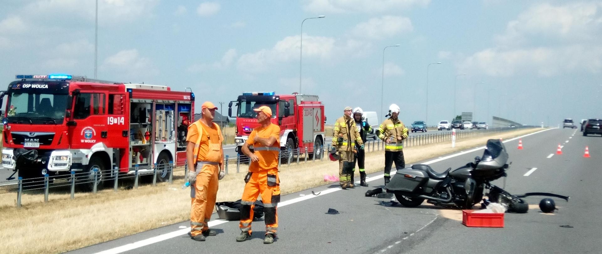 Zdjęcie przedstawia jezdnię na której znajduje się przewrócony motocykl. Wokół pojazdu znajdują się strażacy - w tle widać dwa samochody strażackie.